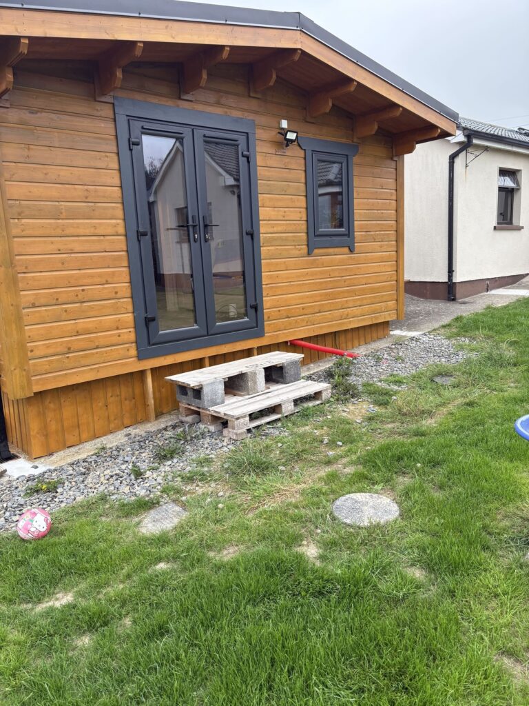 Wooden cabin with black-framed windows, a pallet step, and a grassy area, showcasing Patios Wicklow's outdoor charm