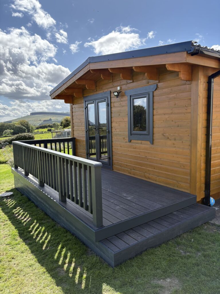 Wooden cabin with a deck, surrounded by green grass and hills under a blue sky with fluffy clouds, showcasing Patios Wicklow