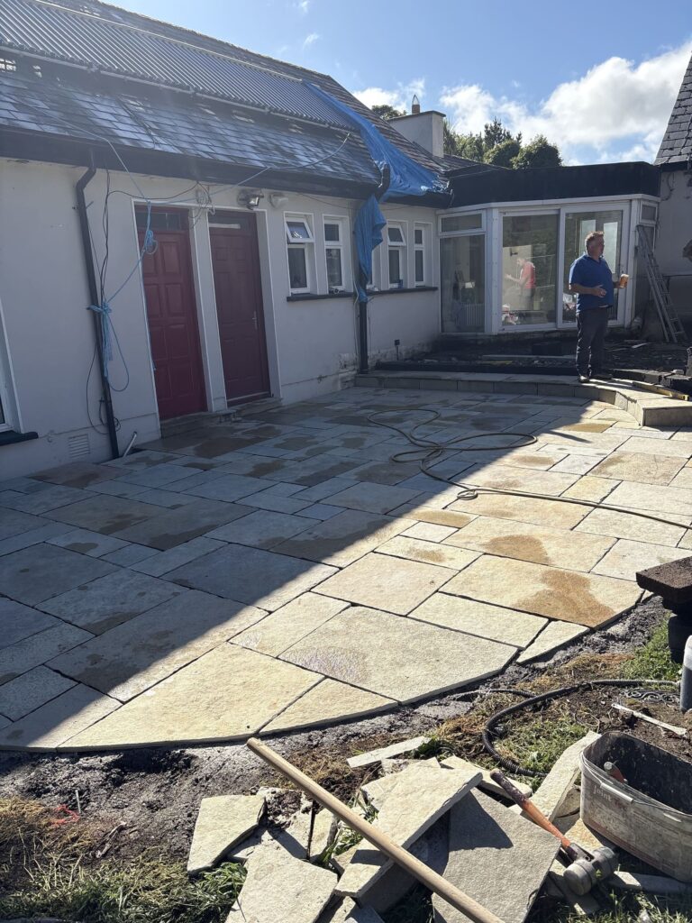 Newly paved sandstone patio in Wicklow, with a worker holding a drink, surrounded by tools and a house with red doors