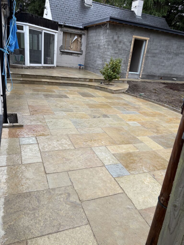 Wet sandstone patio with a newly paved driveway, surrounded by a partially finished house and greenery in Wicklow