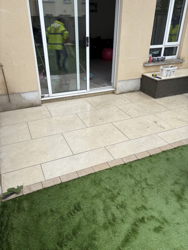 Newly laid patio with light tiles, green artificial grass, and a worker in a reflective jacket near sliding glass doors. Pati