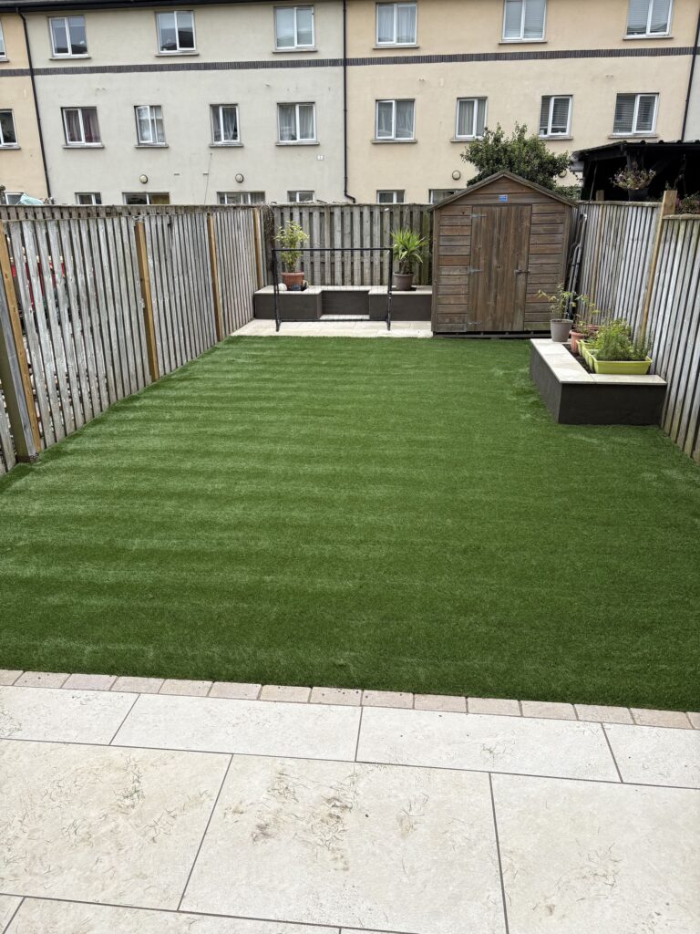 Lush green artificial lawn with stone patio, wooden shed, and planters in a fenced backyard in Wicklow