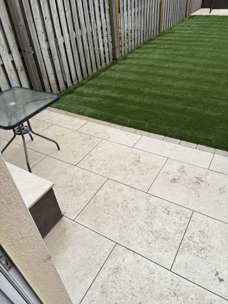 Paved patio area with light stone tiles, a small glass table, and a green artificial lawn in a fenced backyard, Patios Wicklo