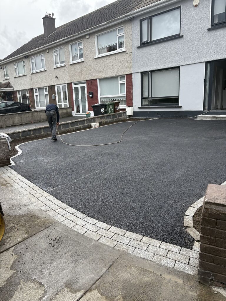 A worker applies resin to a freshly laid driveway, bordered by stone edging, in a residential area with gray houses