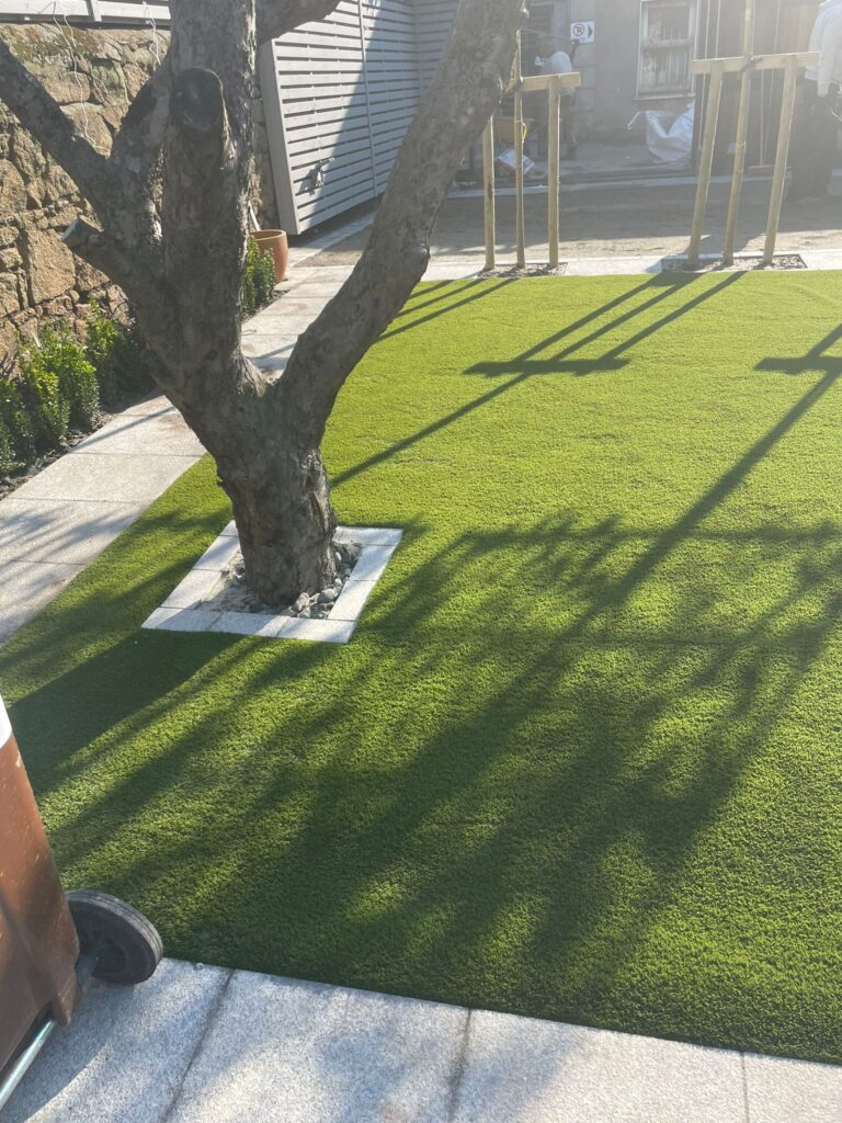 Lush green artificial grass surrounds a tree, with shadows cast on the ground, showcasing Landscaping Wicklow's design