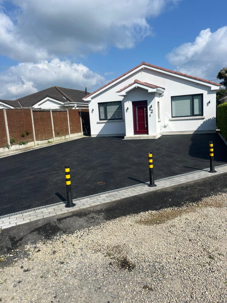 Modern house with a red door, black driveway, and yellow-striped bollards under a blue sky in Wicklow