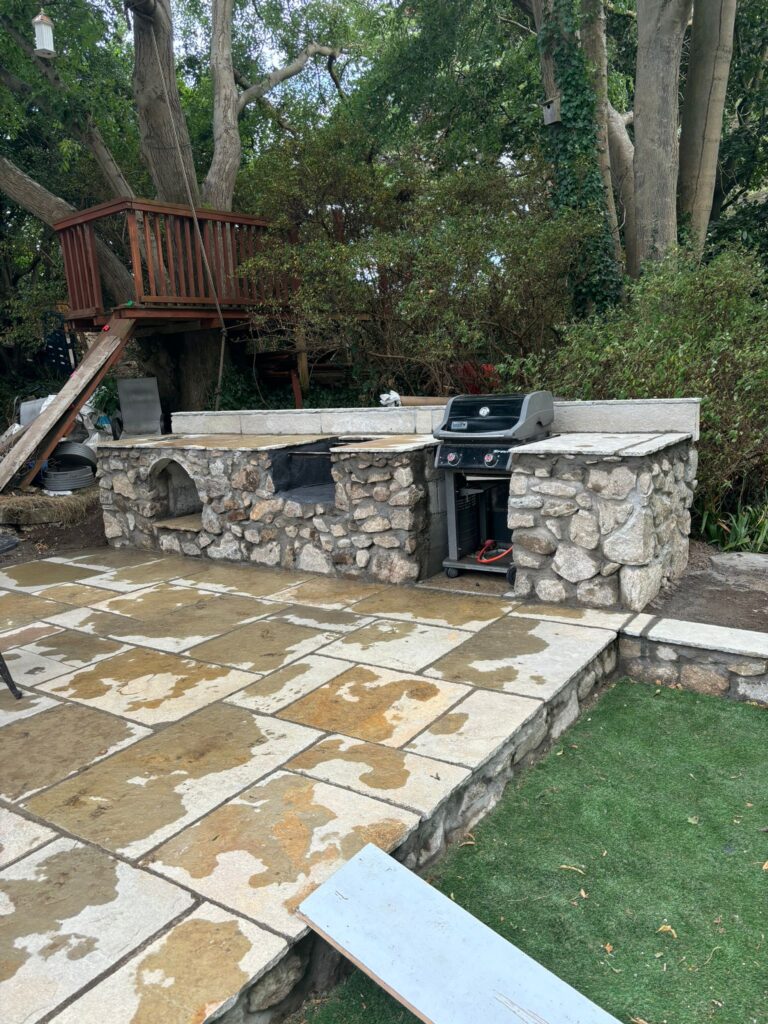Stone patio with a built-in grill, surrounded by greenery and a wooden treehouse in the background, showcasing Patios Wicklow