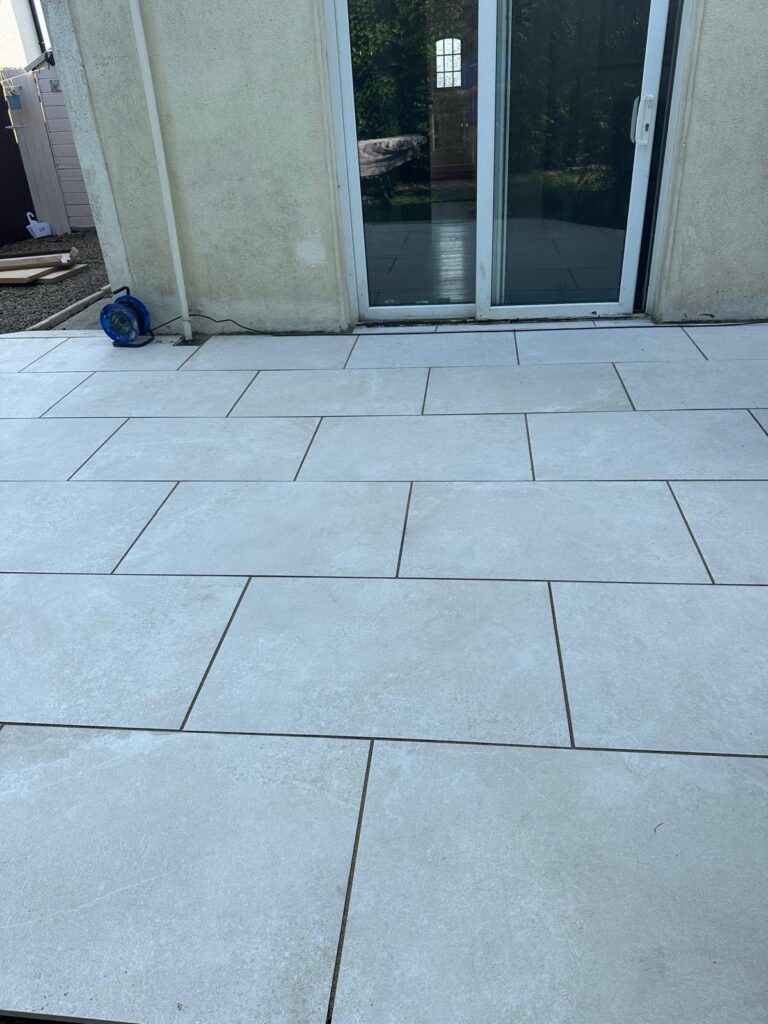 Bright, newly laid light-colored tiles on a patio, leading to a glass door, surrounded by greenery in Patios Wicklow