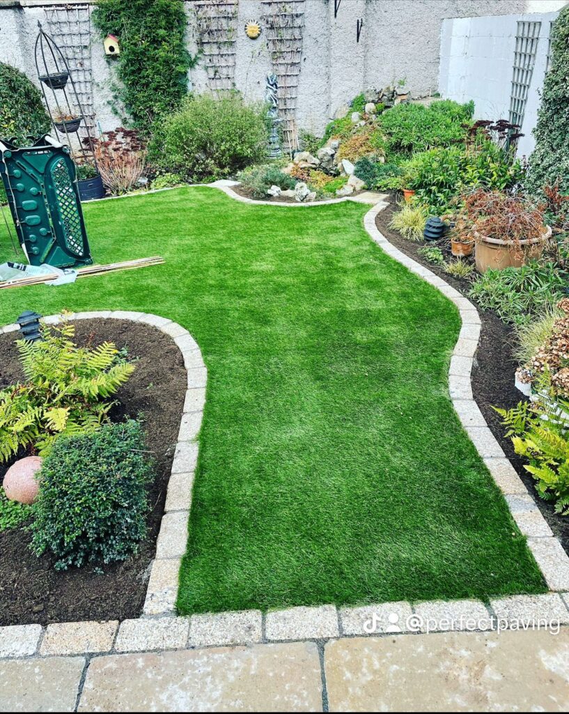 Lush green lawn with curved stone borders, surrounded by various plants and decorative elements in a landscaped garden