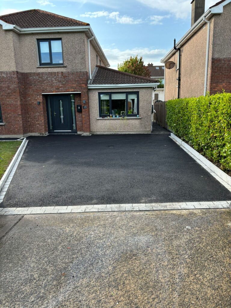 Modern home exterior with a freshly paved driveway, bordered by white stone and green hedges, under a clear blue sky