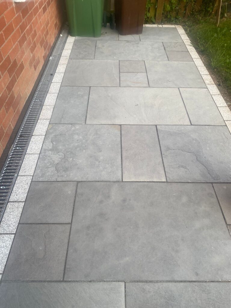 Gray sandstone patio with a drainage channel, bordered by light stones, and green and brown bins nearby in a garden setting