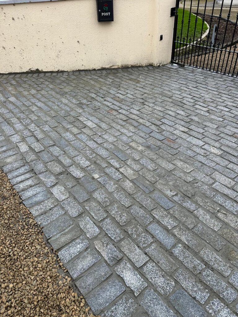 Paved driveway with gray cobblestones, bordered by gravel, next to a wall with a black mailbox labeled "POST"