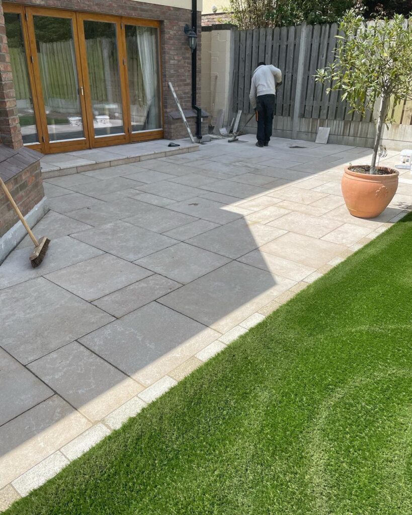 A worker lays gray paving stones on a patio beside a lush green lawn and a terracotta pot with a small tree in Wicklow