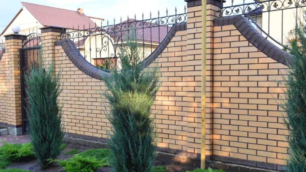 Brick wall with decorative ironwork, flanked by tall green shrubs, leading to a driveway in a residential setting in Wicklow