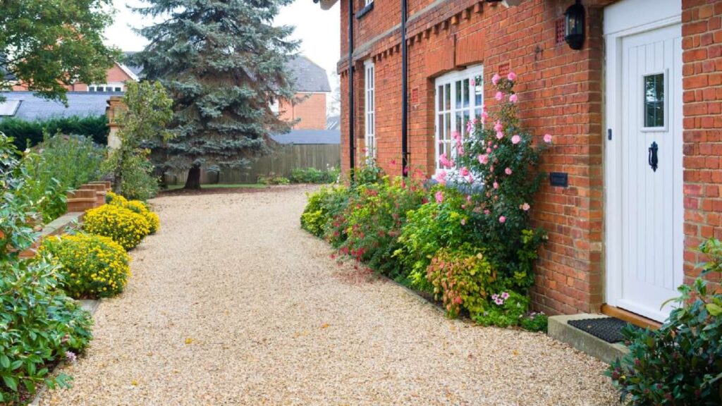 Gravel driveway lined with colorful flowers and shrubs, leading to a brick house with a white door in a serene setting