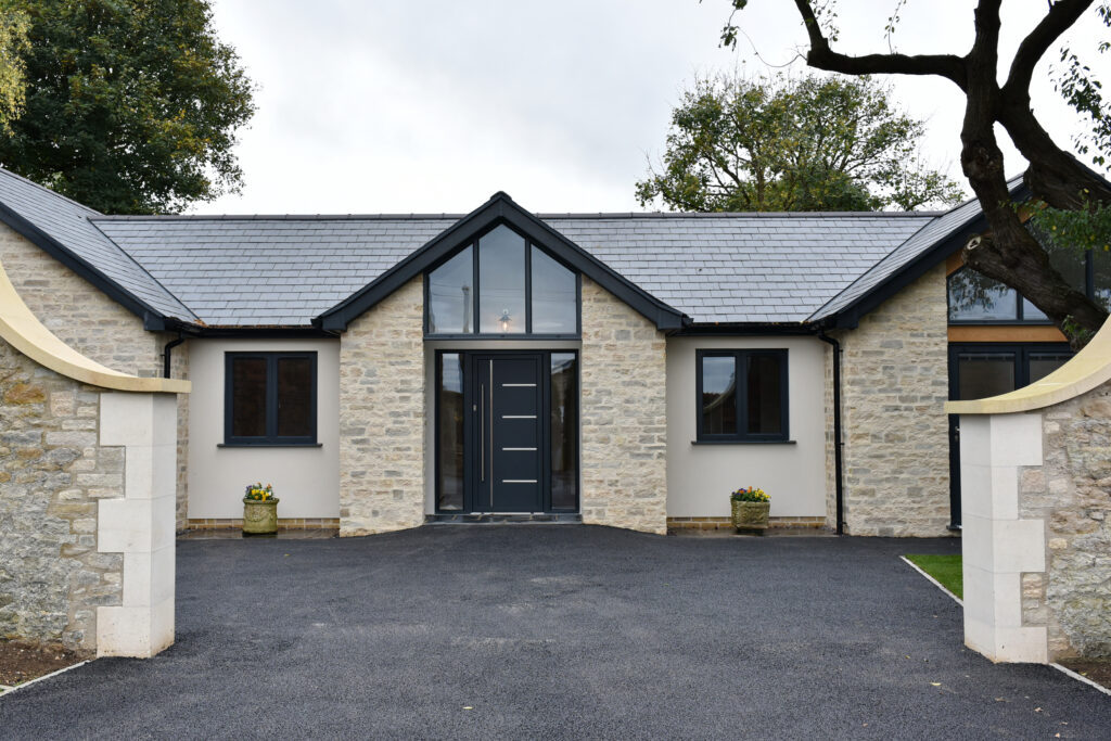 Modern stone house with a dark door, surrounded by a black asphalt driveway and green planters, set in a tranquil environment