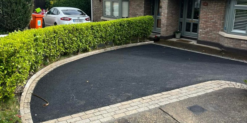Driveway Contractors Dublin And Wicklow. Curved driveway with a stone border, leading to a house with a brick facade and green hedges in a residential area.