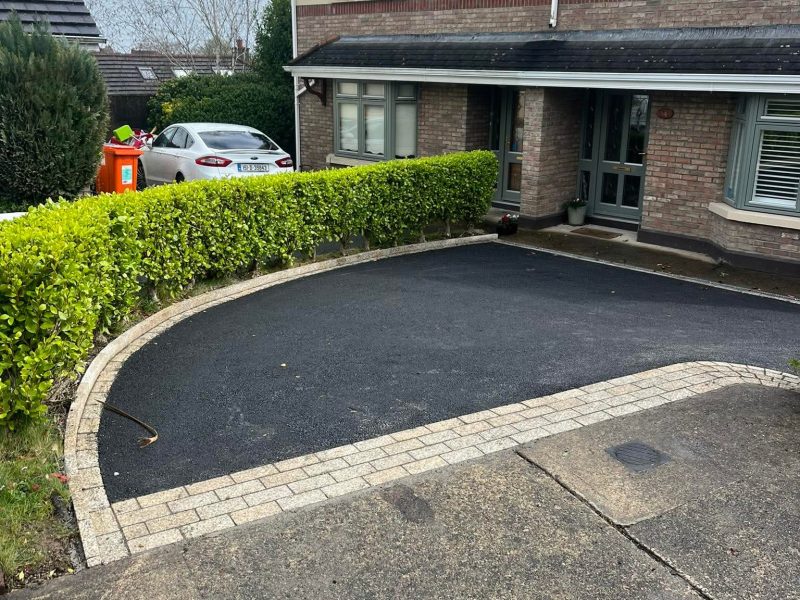 Driveway Contractors Dublin And Wicklow. Curved driveway with a stone border, leading to a house with a brick facade and green hedges in a residential area.